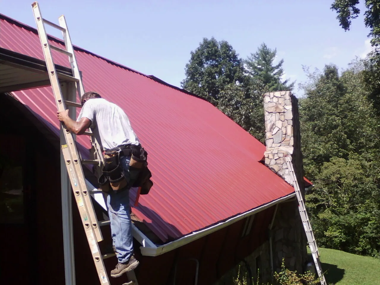 Roofer on a ladder beside a red metal roof and stone chimney — Soffit & Fascia Repair by Roofing Brothers in Millmont